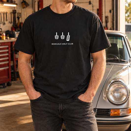 Styled mockup of the Black Essential tee worn beside a silver sports car, showing the white pedal artwork