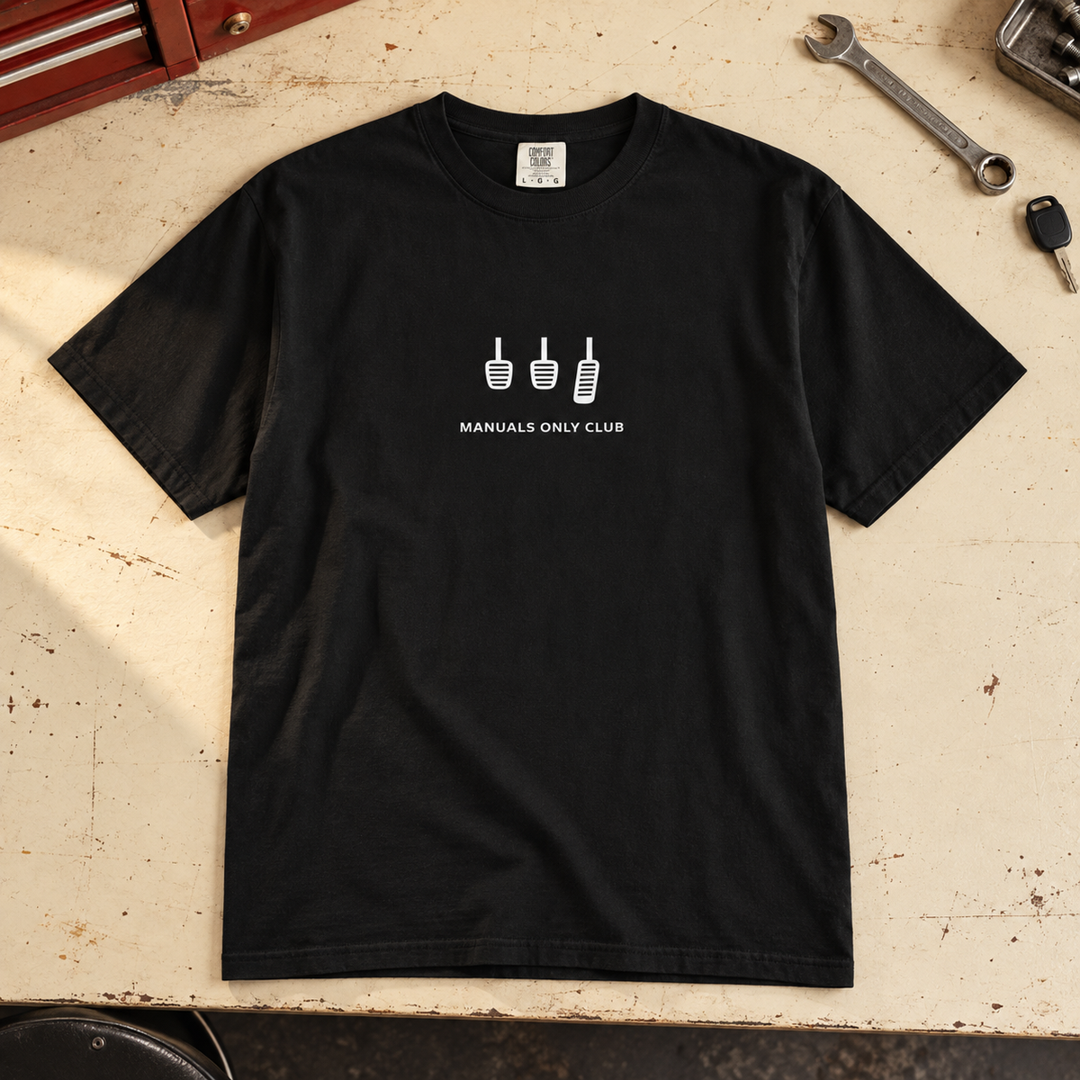 Styled mockup of the Black Essential tee with white pedal artwork on a garage workbench