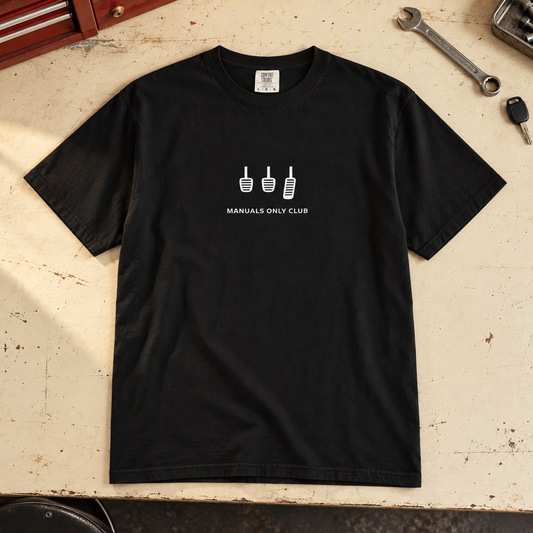 Styled mockup of the Black Essential tee with white pedal artwork on a garage workbench